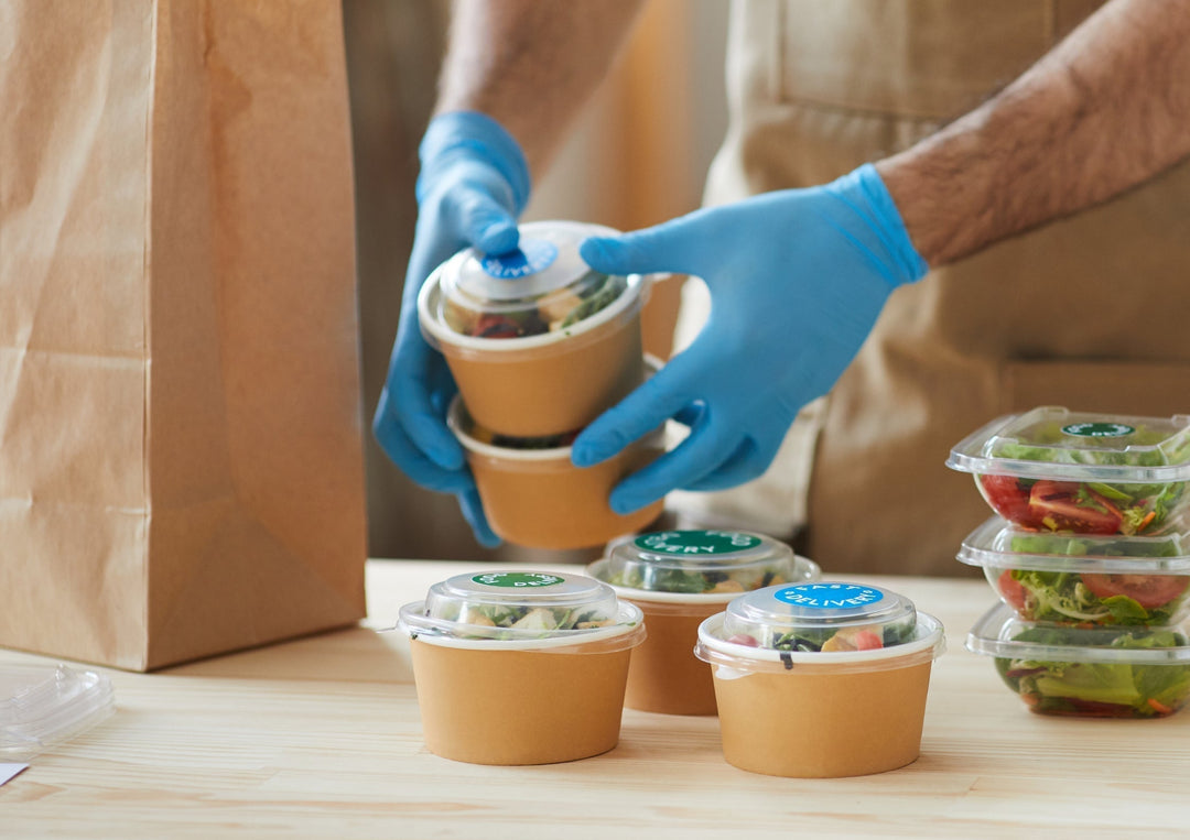comptoir de cuisine en plein rush, avec plusieurs commandes en cours de préparation, chaque repas étant identifié dans une boîte kraft ou bagasse avec une étiquette ou une inscription visible